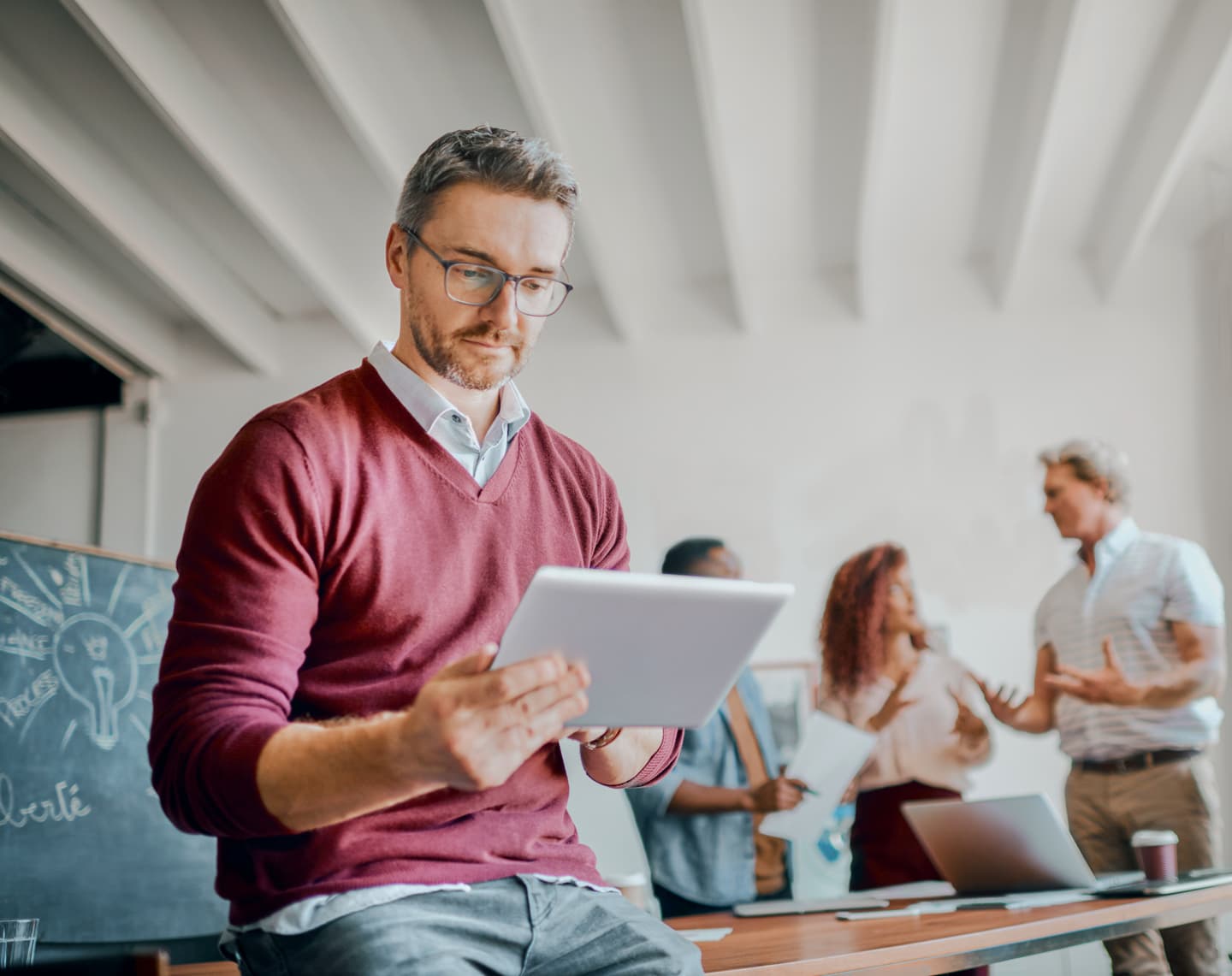 Person in burgundy jumper using tablet in modern office with colleagues discussing in background.