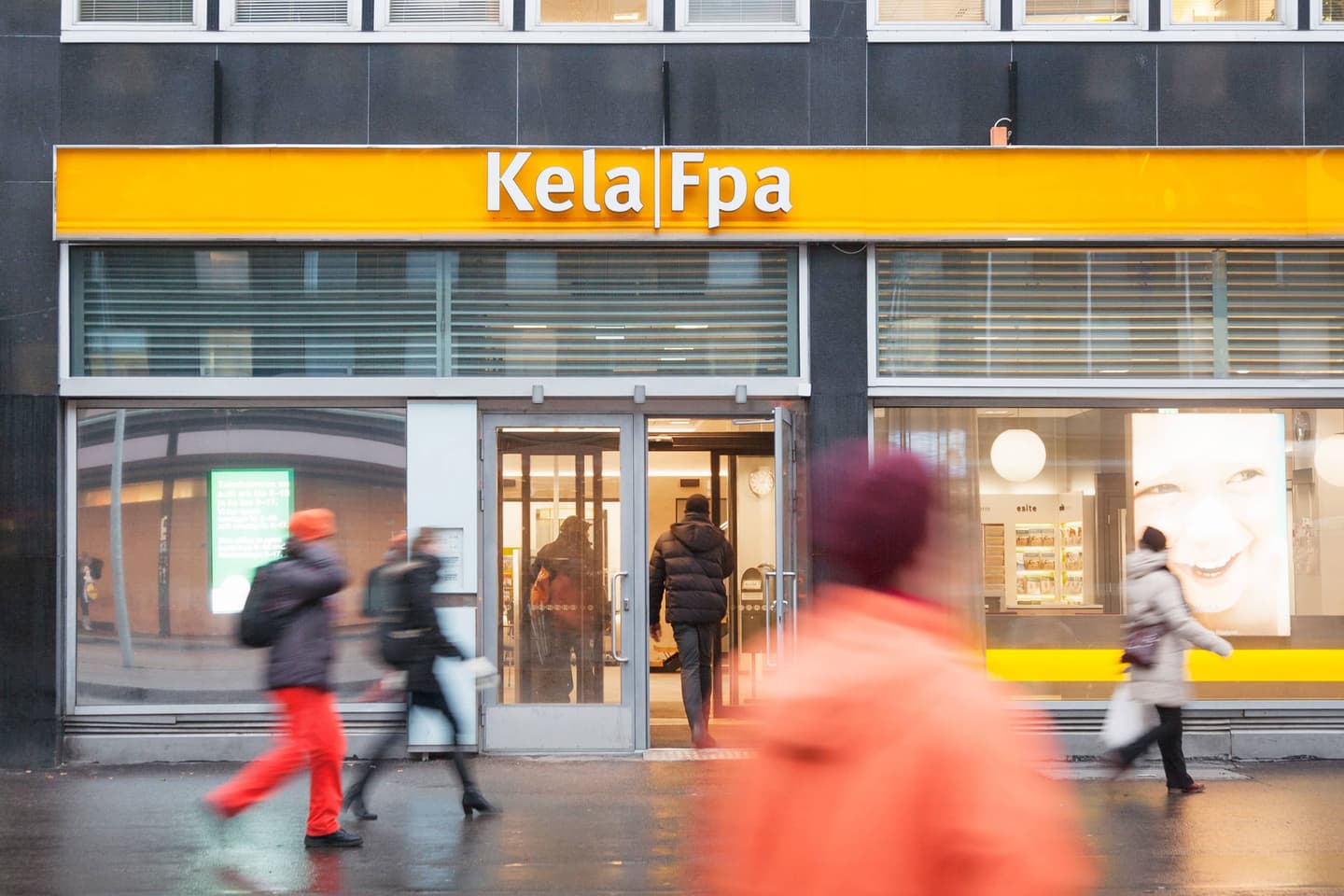 Kela|Fpa office storefront with bright yellow sign, glass entrance doors, and blurred pedestrians walking past in winter clothing.